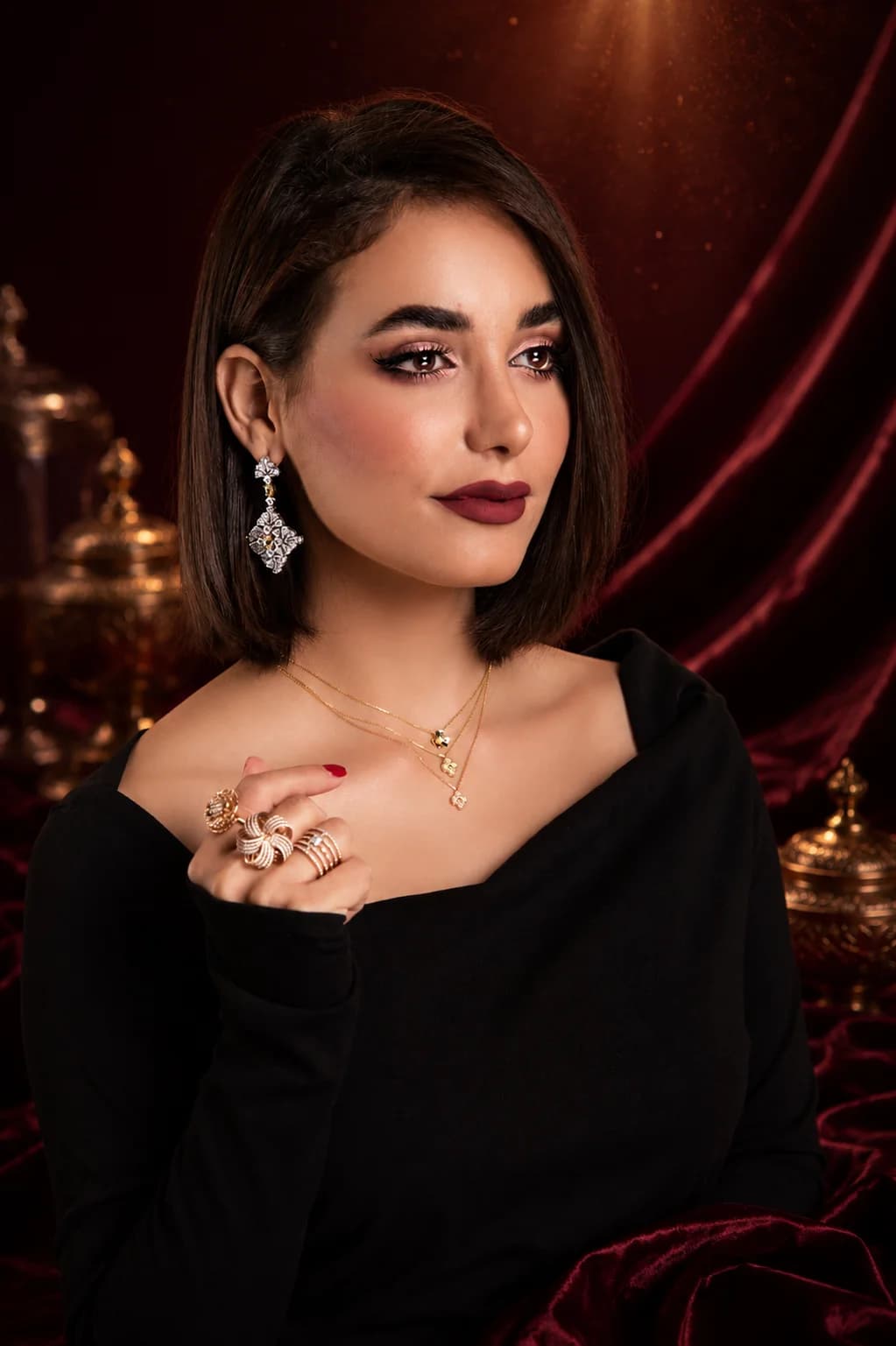 Model wearing statement earrings, layered necklaces, and rings against a maroon velvet backdrop